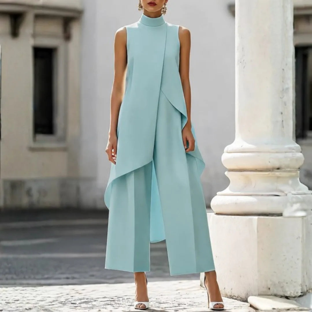 Woman wearing an ice-blue sleeveless jumpsuit with high neck and flowing asymmetric drape over wide-leg trousers.