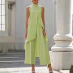 Sleeveless green jumpsuit with high mock neck and layered asymmetric front, shown on a stone pathway.