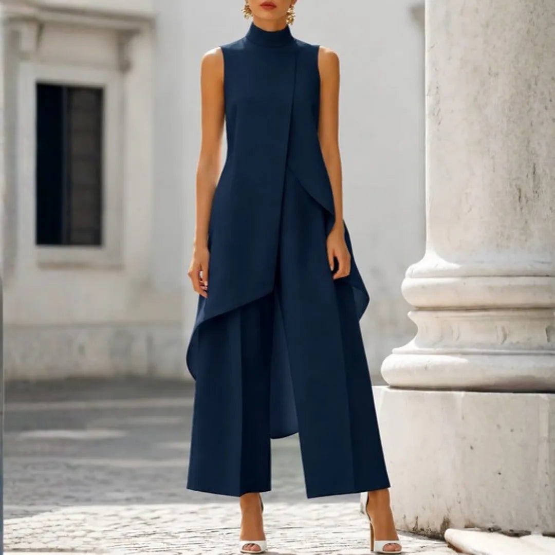 Navy sleeveless jumpsuit with draped asymmetric panel and wide culotte-style legs, worn with strappy heels.