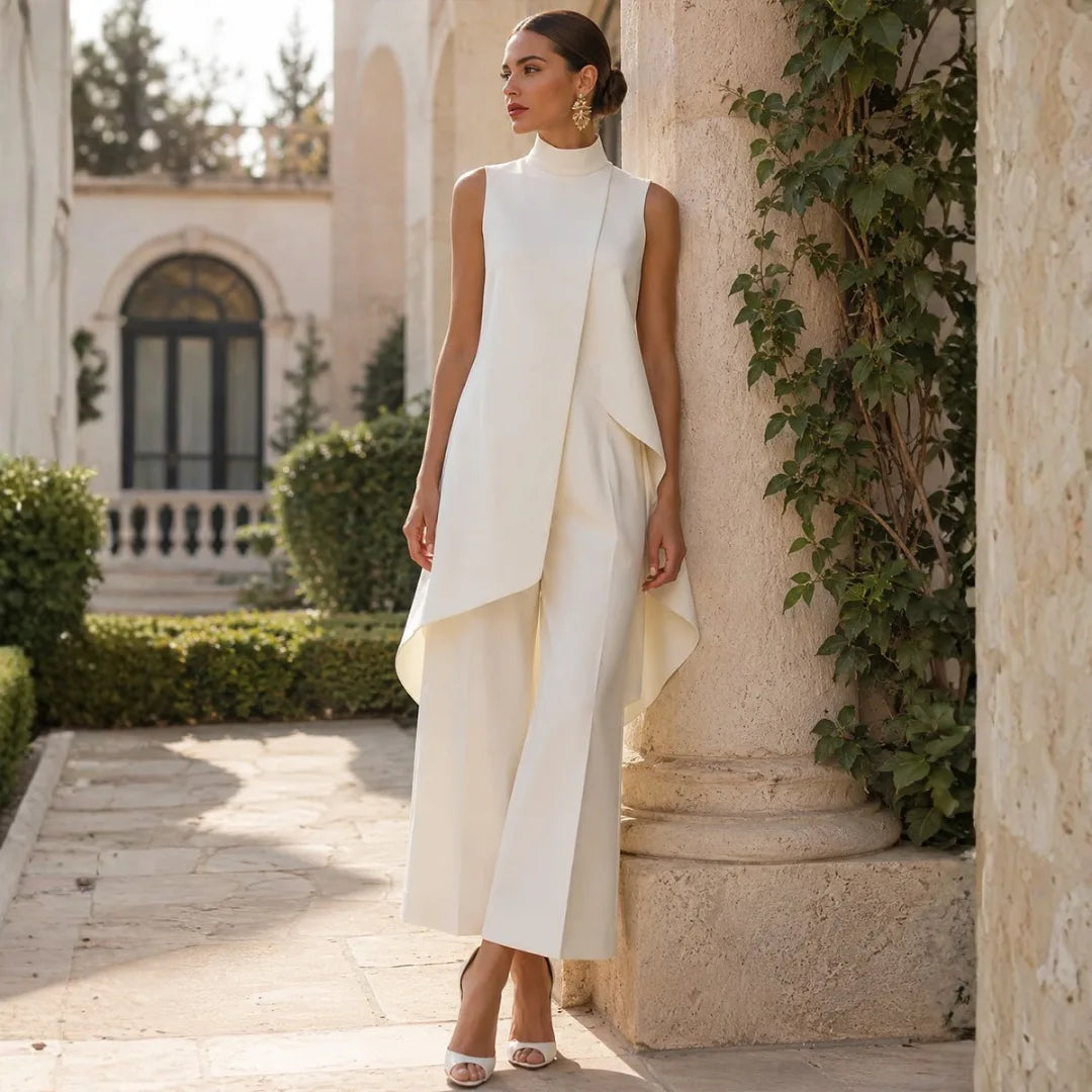 Ivory jumpsuit worn leaning against a stone pillar, showing the high neckline, draped front and wide cropped legs.