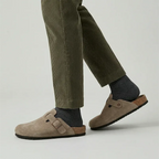 Close-up of a person wearing beige suede buckle clogs with grey socks and olive corduroy pants, shown mid-step.