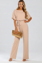 Model wearing a beige jumpsuit with boat neckline, tied waist belt and wide-leg trousers, holding a gold clutch