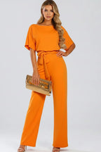 Orange jumpsuit with boat neck, relaxed short sleeves and a waist tie, styled with a metallic gold clutch