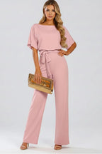 Model in a blush pink jumpsuit with draped sleeves, belted waist and wide-leg trousers, holding a gold clutch