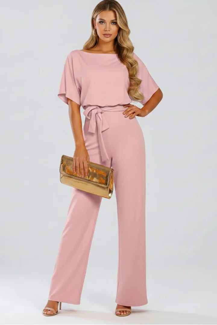 Model in a blush pink jumpsuit with draped sleeves, belted waist and wide-leg trousers, holding a gold clutch