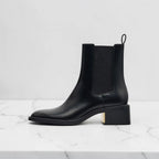 Side view of black leather Chelsea boot with pull tab, elastic gusset, and low block heel with gold accent.