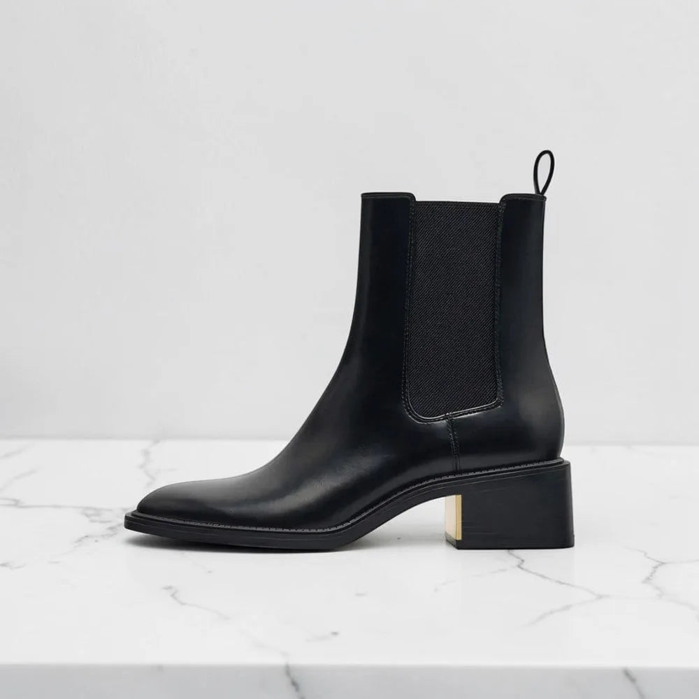 Side view of black leather Chelsea boot with pull tab, elastic gusset, and low block heel with gold accent.
