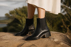 Woman wearing black leather Chelsea boots with a white flowing dress while standing on a rock outdoors.