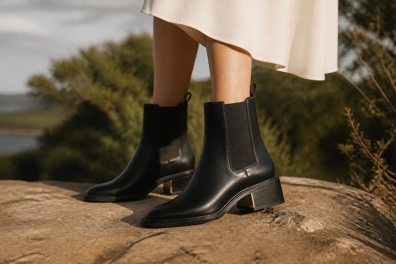 Woman wearing black leather Chelsea boots with a white flowing dress while standing on a rock outdoors.