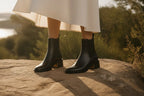Close-up of black Chelsea boots paired with a white dress, showing the square toe and heel detail in sunlight.