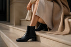 Black leather Chelsea boot shown on stone steps next to a seated woman in a beige coat, highlighting the heel design.