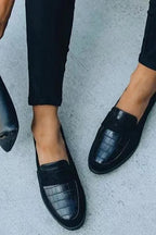 Close-up of black croc-embossed leather penny loafers worn with cropped black pants, angled front view on concrete.