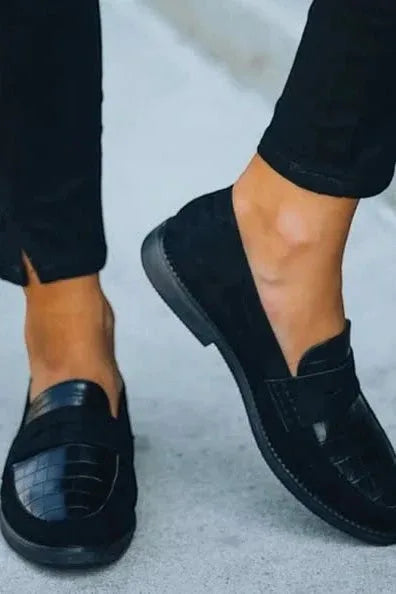 Close-up of the loafer's low stacked sole and suede upper paired with black trousers, standing on grey pavement.