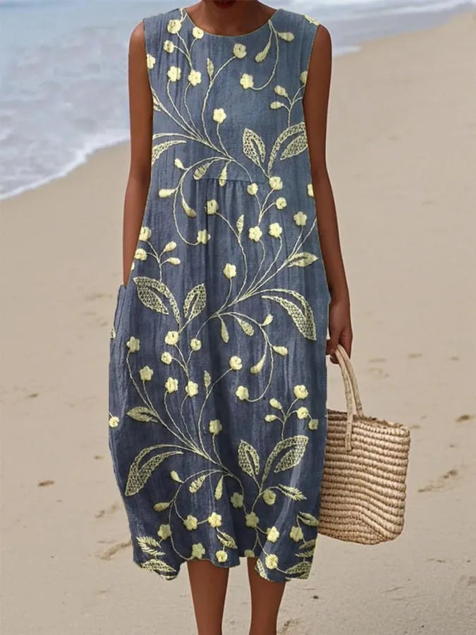 Sleeveless denim-blue midi dress with yellow embroidered floral vine design, worn on a sandy beach.