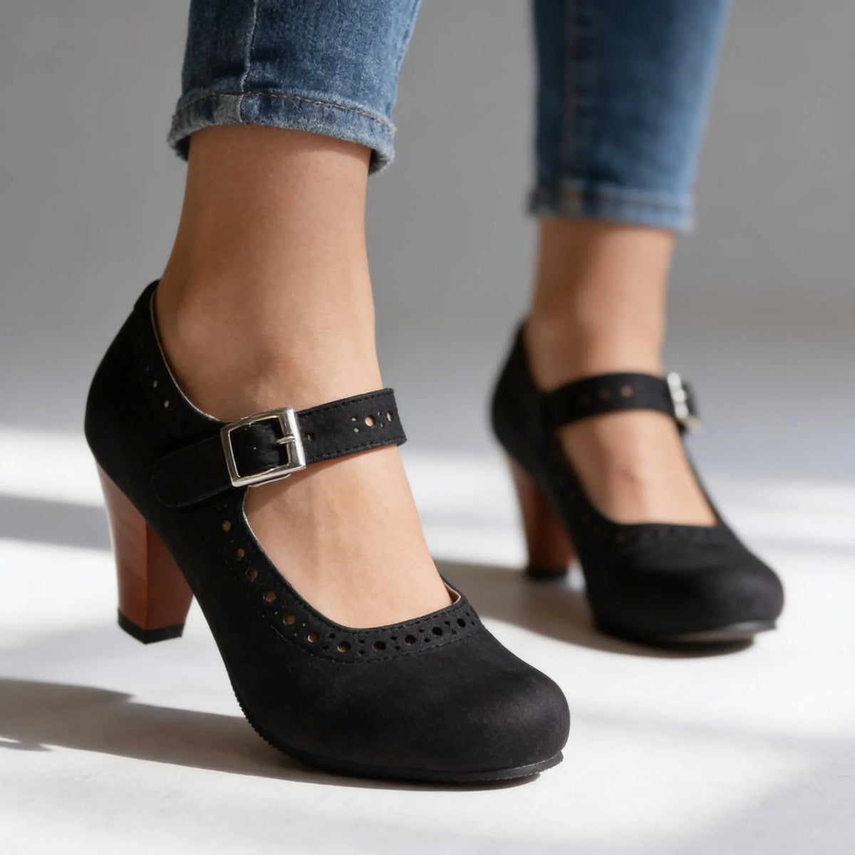Black suede Mary Jane heels on a plain studio backdrop, showing the rounded toe and ankle strap buckle