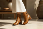 Woman in motion wearing camel leather ankle boots with a ribbed cream skirt, highlighting the round toe and heel height