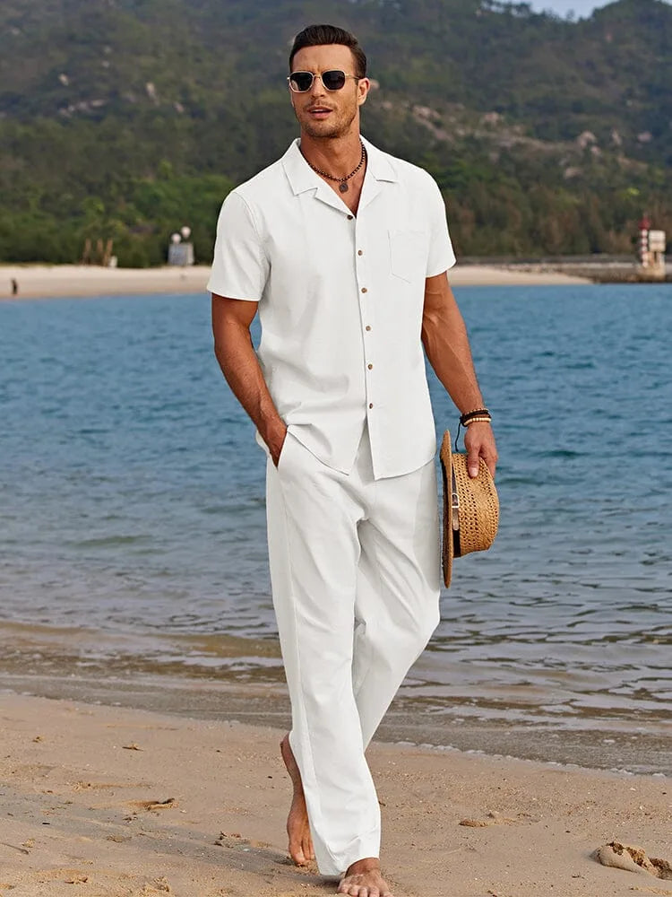 Man in white camp collar shirt and matching trousers walking barefoot along the beach, holding a straw hat.