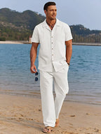 Man walking on the shoreline in a white camp collar shirt and trousers set, holding sunglasses in hand.