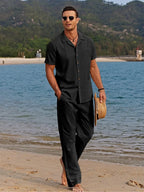 Man strolling on sand in a black camp collar shirt and matching trousers, carrying a straw hat.