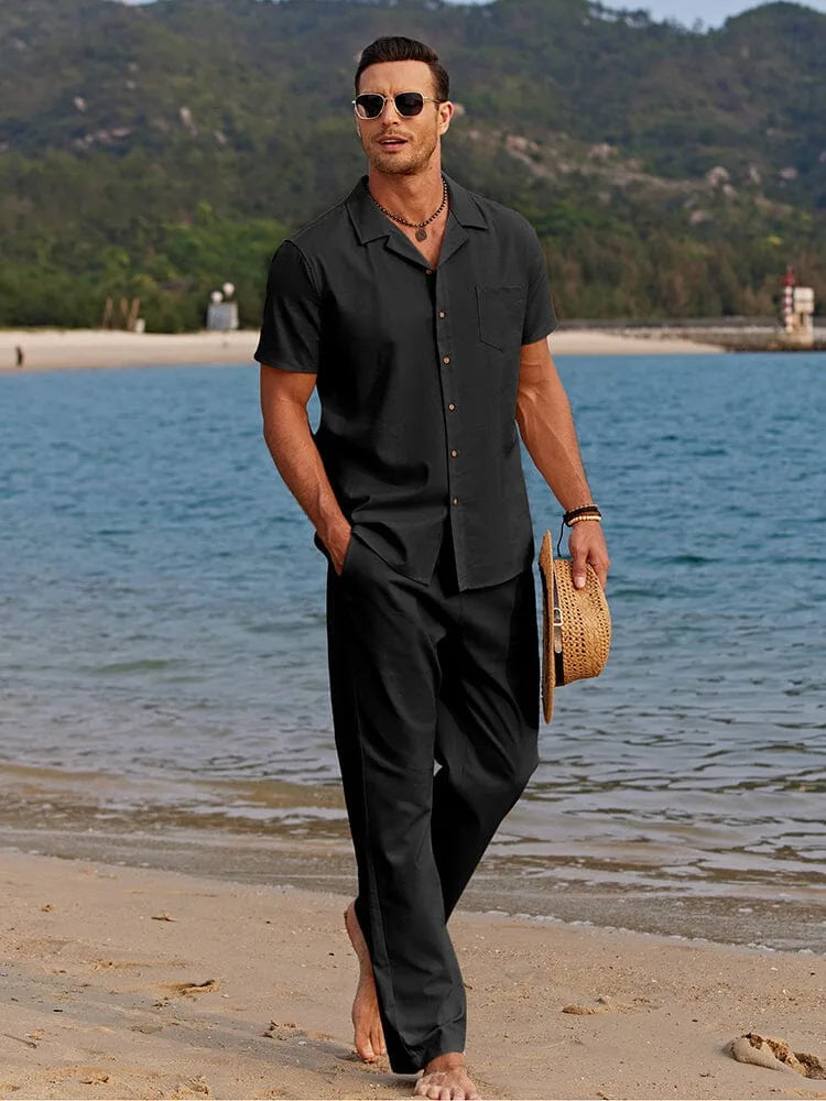 Man strolling on sand in a black camp collar shirt and matching trousers, carrying a straw hat.