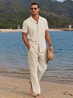 Man walking along the beach in a cream camp collar shirt and trousers set, holding a straw hat.