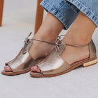 Model wearing champagne leather sandals with denim jeans, showing bow-tie lace closure across the foot