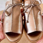 Close-up of the bow-tie lace detail and stitched leather upper on a pair of champagne metallic sandals