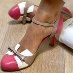 Front view of pink and beige colour-block heels with ankle strap and bow, worn with a white handbag nearby