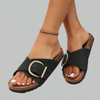 Close-up of black faux-leather cork-footbed slide sandals with oversized antique-gold buckle, worn on crossed feet.