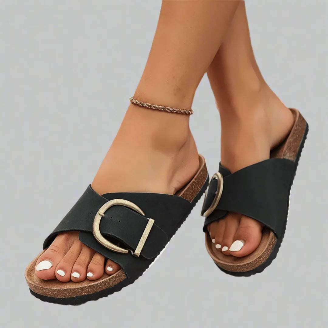 Close-up of black faux-leather cork-footbed slide sandals with oversized antique-gold buckle, worn on crossed feet.