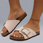 Pale cream cork-footbed slide sandals with brushed gold buckle detail, shown from the side on bare feet.