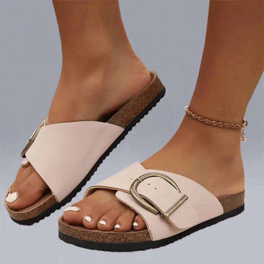 Pale cream cork-footbed slide sandals with brushed gold buckle detail, shown from the side on bare feet.