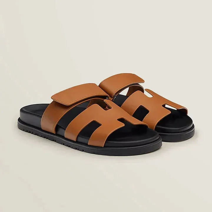 Tan leather slide sandals with cut-out H-strap design and chunky black sole, shown at an angle.