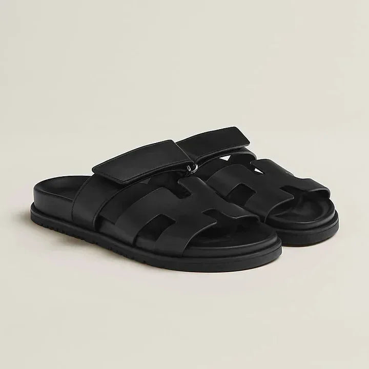 Black leather slide sandals with wide adjustable strap and cut-out panel detail on a black sole.