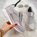 Hand holding one grey and silver sneaker beside its matching pair on a fluffy white rug