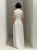 Back view showing the sleeveless maxi dress with stand collar and belted waistband against a minimalist plaster wall