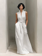 Full-length front view of the white sleeveless maxi dress styled with a loosely knotted belt in a sunlit stone corridor