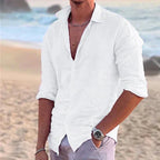 Man wearing a white cotton linen shirt open at the collar, standing on a sandy beach near the ocean