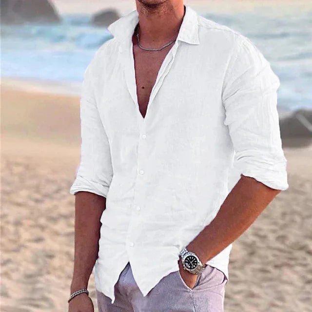 Man wearing a white cotton linen shirt open at the collar, standing on a sandy beach near the ocean