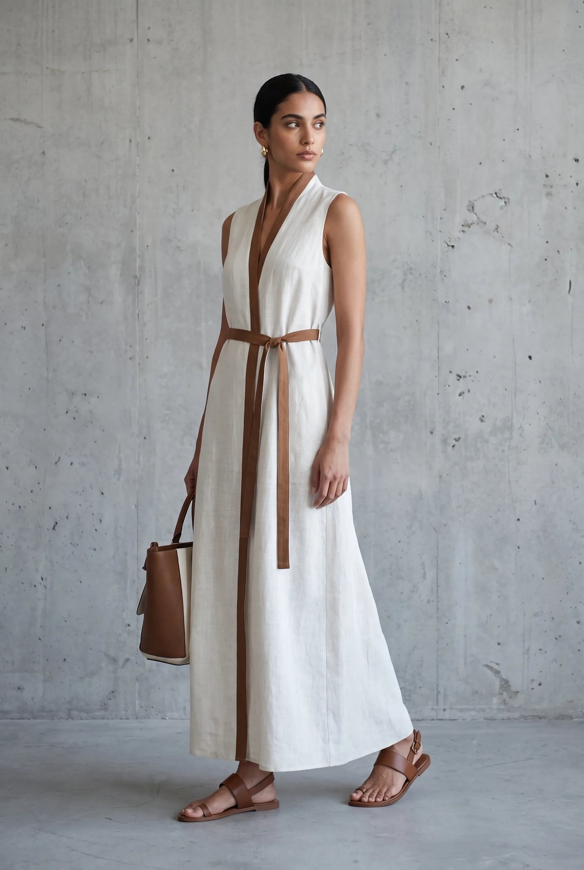 Woman walking in a sleeveless cream linen maxi dress with tan leather trim and tie belt, carrying a brown tote bag