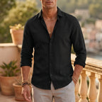 Man in a black cotton linen shirt, fully buttoned, paired with light grey trousers and layered bracelets outdoors.