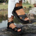 Black crossover strap platform sandal with ankle buckle closure, worn outdoors on a rock near a lake