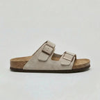 Side view of a light beige suede double-strap sandal with bronze buckles and brown outsole