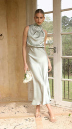 Front view of model in sage green satin midi dress with draped high collar and bouquet, standing by glass doors