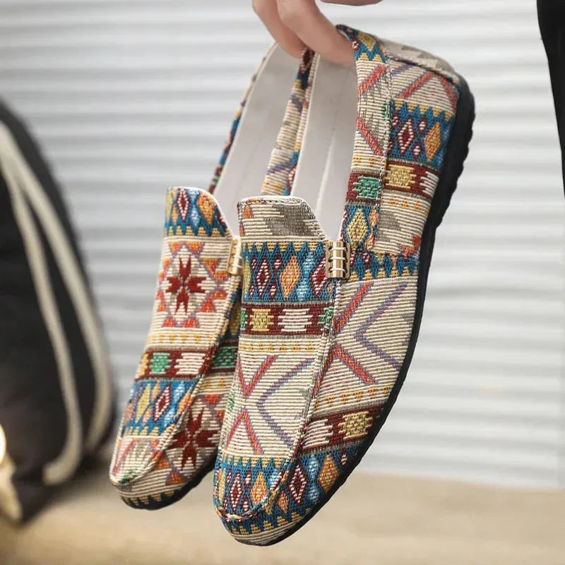 Hand holding a pair of beige ethnic-pattern woven loafers, showing gold hardware detail and padded white collar