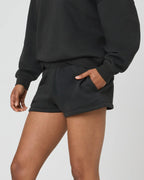 Close-up side view of woman wearing black sweat shorts with hand in side pocket, paired with oversized black jumper