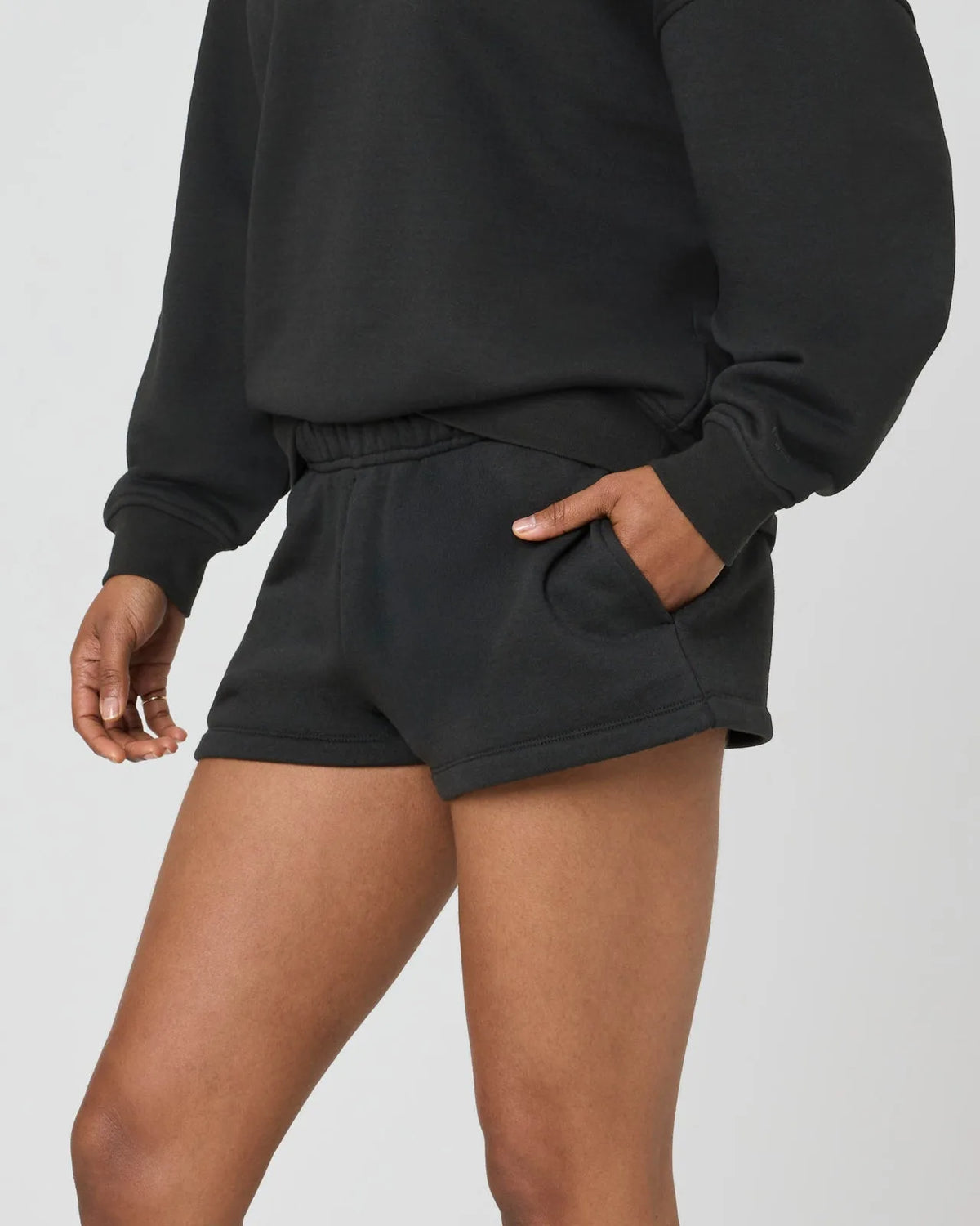 Close-up side view of woman wearing black sweat shorts with hand in side pocket, paired with oversized black jumper