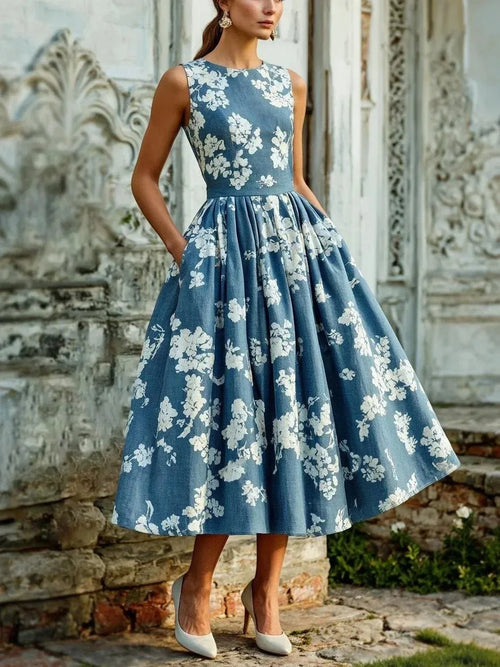 Woman in sleeveless denim-blue floral midi dress with full pleated skirt, hands in side pockets, standing by ornate stone wall