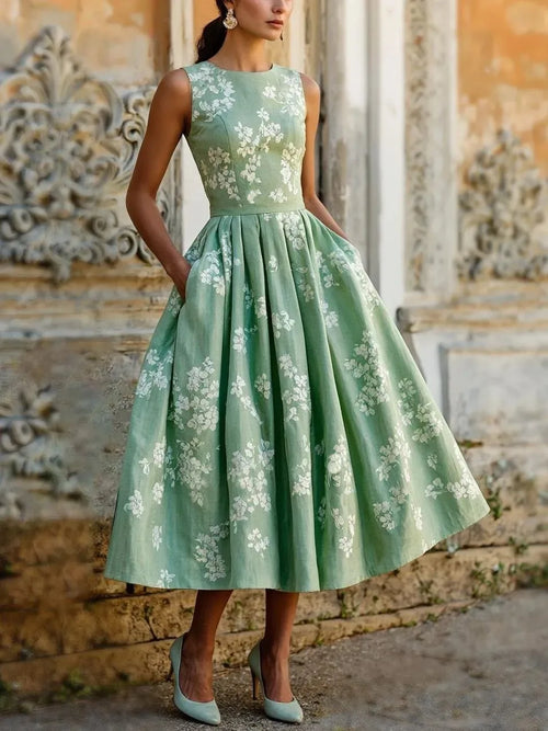 Front view of sage green floral sleeveless midi dress with fitted bodice and flared pleated skirt against weathered stone facade