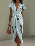 Front view of a light blue wrap midi dress with cap sleeves, waist tie and printed leaf motif against a plain backdrop